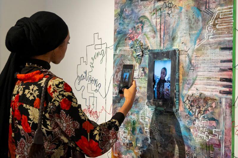 Young woman holding up her phone to a multi-media work hanging on the wall of the Mason Gross Galleries