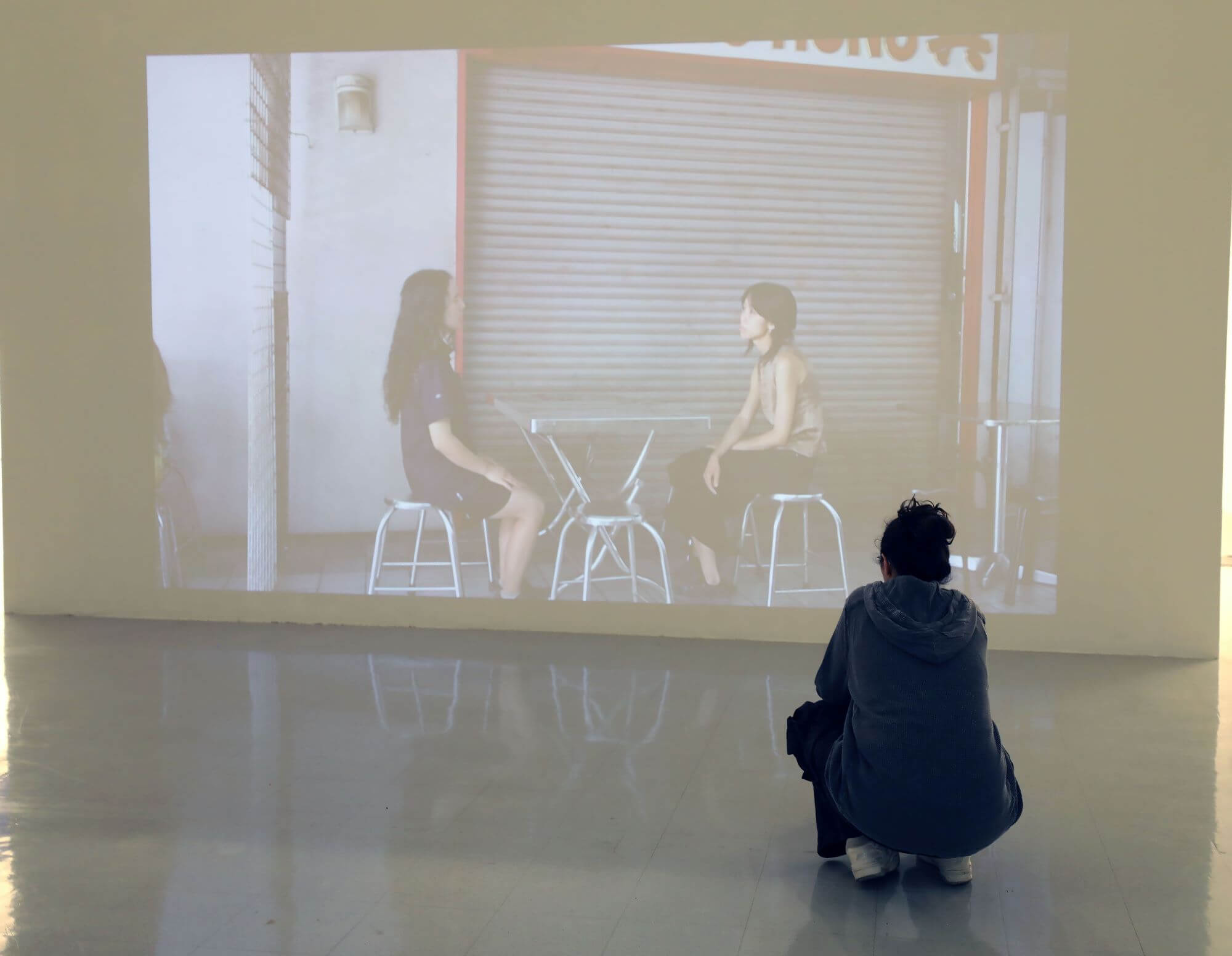 Person crouching in the galleries in front of a screen projecting an image of two people at a small table.