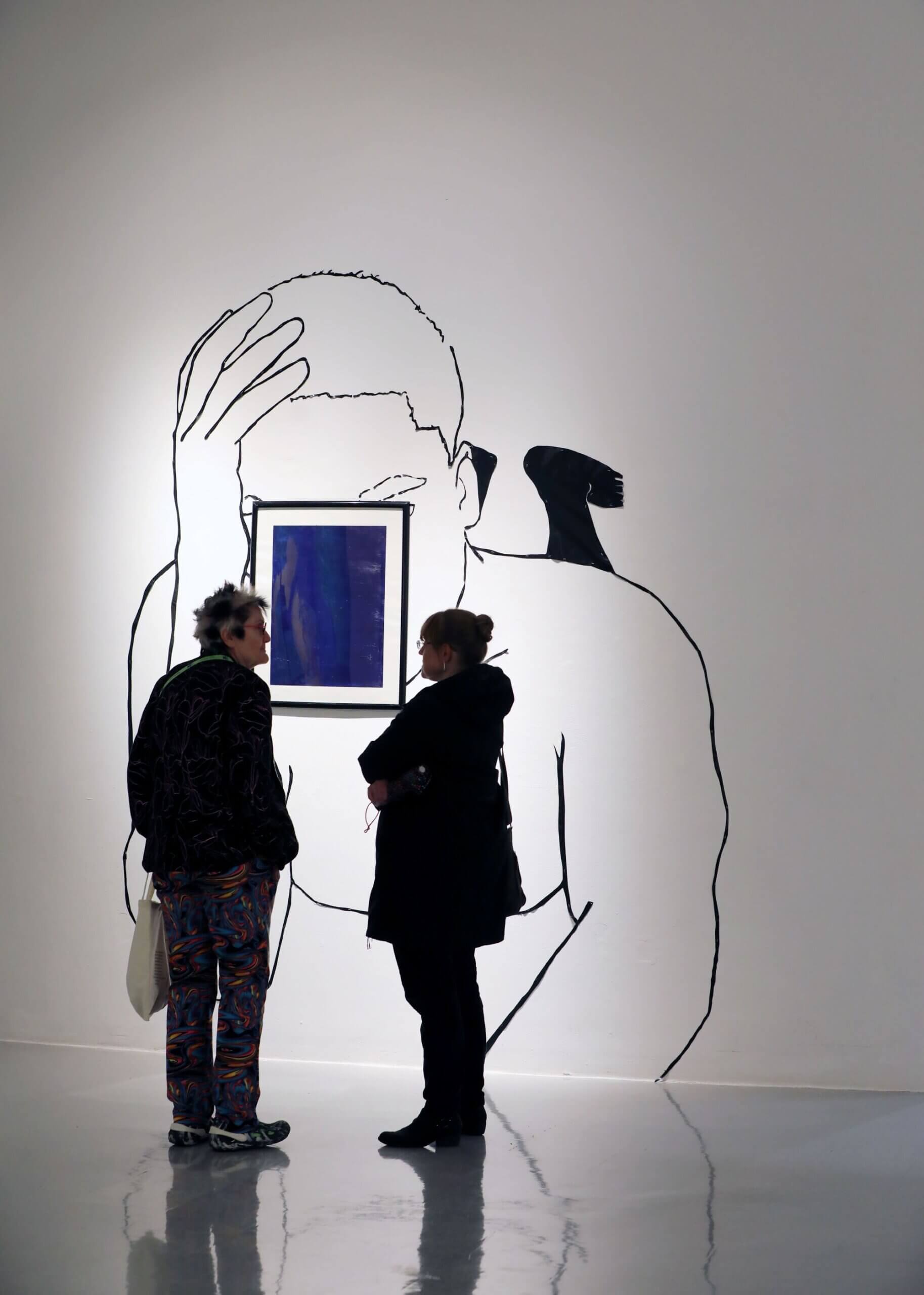 Two people in the gallery looking at a work painted on the wall with a mirror where the face would usually be.