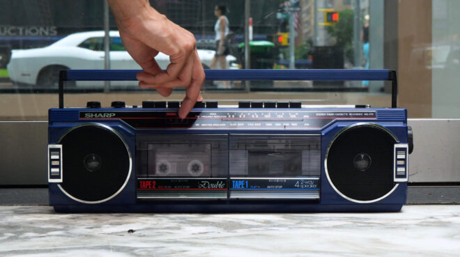 A hand is reaching down to click a button on an old school stereo that sits on the ground.