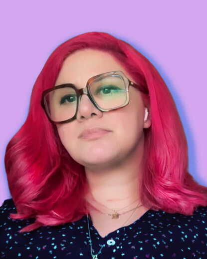 A photo of Emily Sara. She has flaming pink medium-length hair, oversized square glasses and a black dress with tiny red and white flowers with a blue cast over it. She is wearing a few gold necklaces and a scar across her throat is visible. The background is light purple with a dark blue drop shadow.