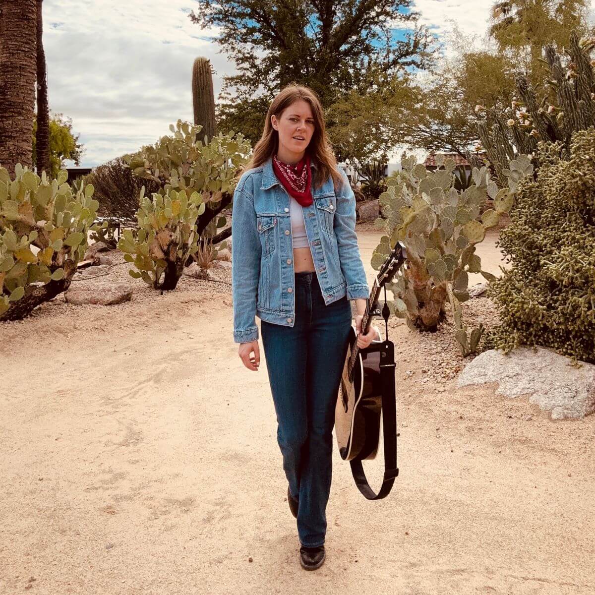 Woman in denim jacket with a red bandana around neck carries acoustic guitar in desert.